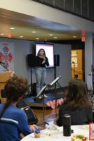 A 12th Grade student stands on a stage and addresses the room as other students look on