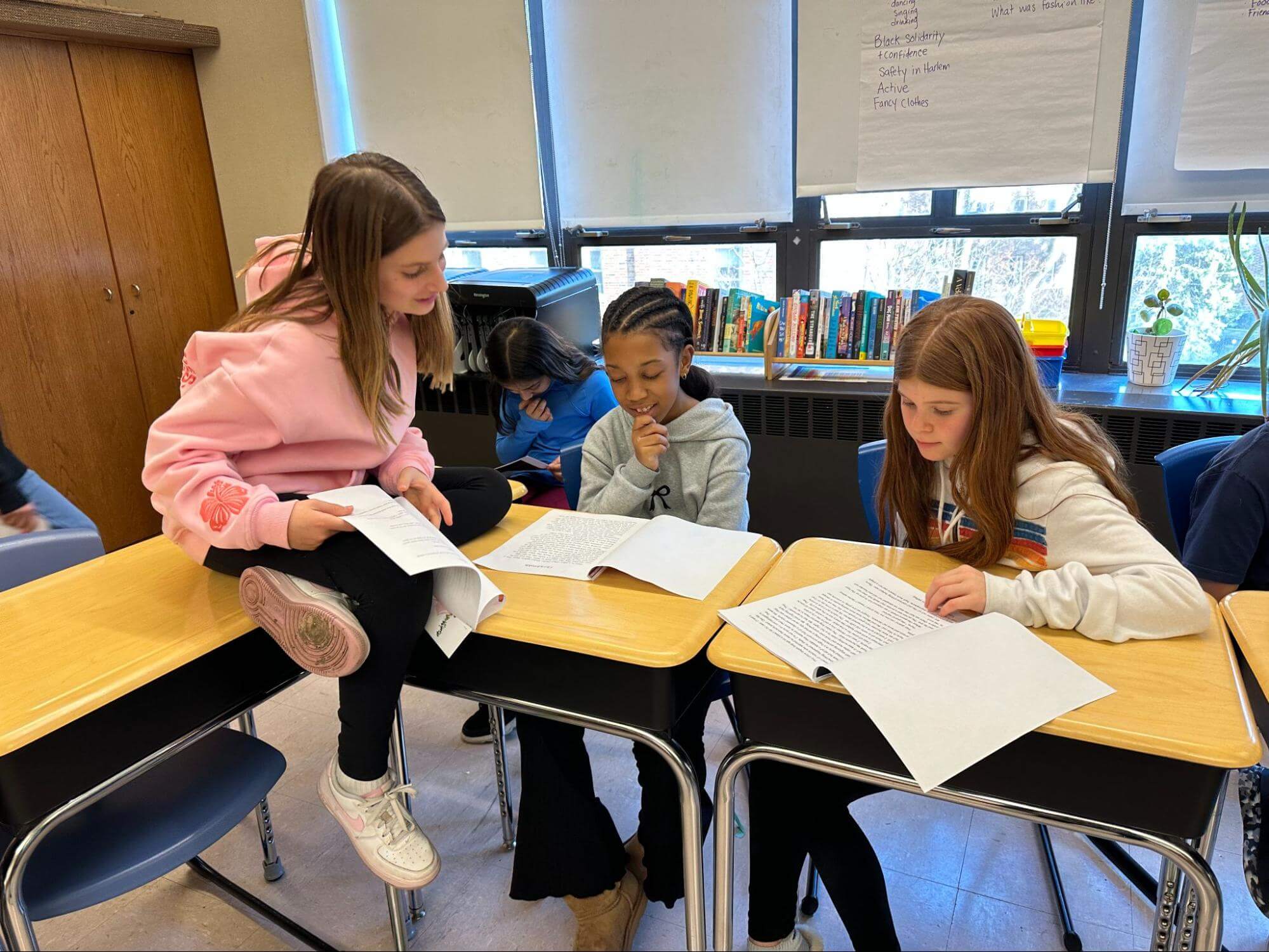 Three Ethical Culture Fieldston School Fieldston Lower students sit at a desk smiling as they look down at the stories they are reading.