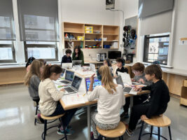 Students sit around collaborative desk in classroom and work on Scratch coding project.