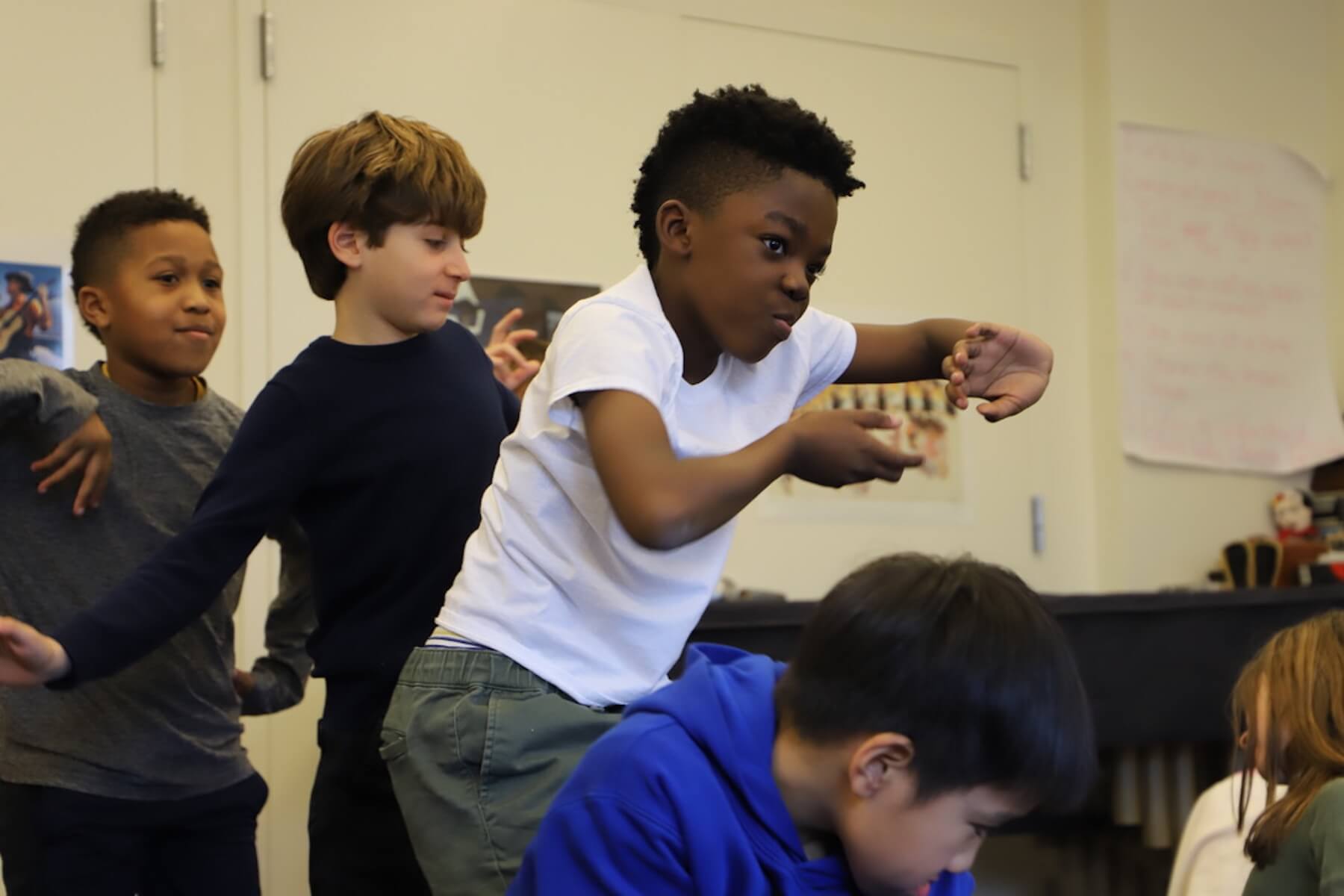 Three Fieldston Lower 3rd Graders stand in a line as they practice their dance moves for the musical. They are all making expressive gestures with their faces and hands.