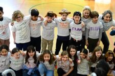 Fieldston Lower 3rd Graders stand with their arms around each other, smiling and singing brightly.