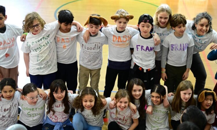 Fieldston Lower 3rd Graders stand with their arms around each other, smiling and singing brightly.