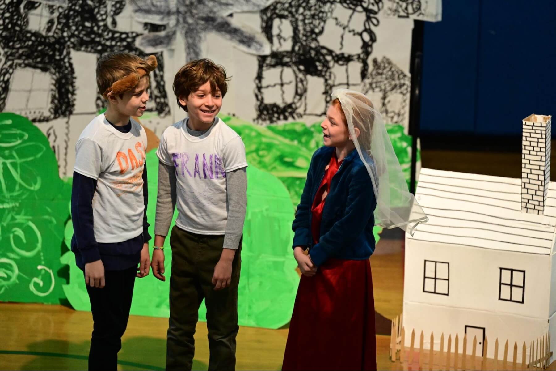 During a performance of the 3rd Grade musical, three students stand smiling in front of a green set.