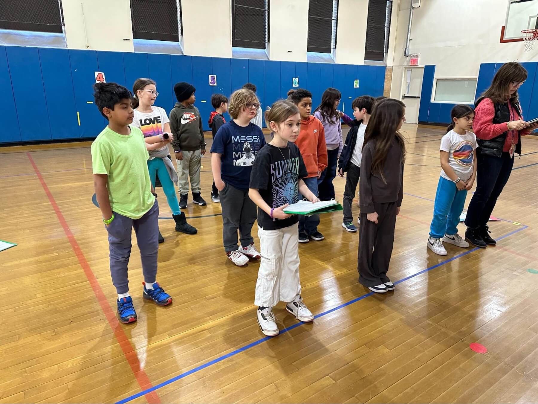 A large group of Fieldston Lower students stands in a crowd, rehearsing a piece of blocking for the 3rd grade musical. Some students hold scripts in their hands.