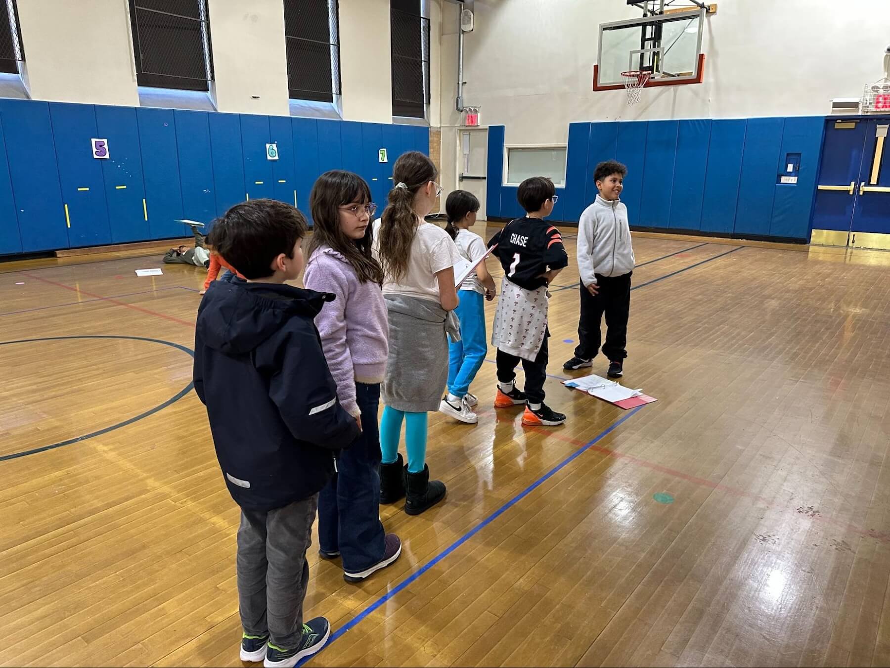 Several Fieldston Lower 3rd Graders stand in a line and look intently at a student delivering a line in rehearsals for the 3rd Grade musical.