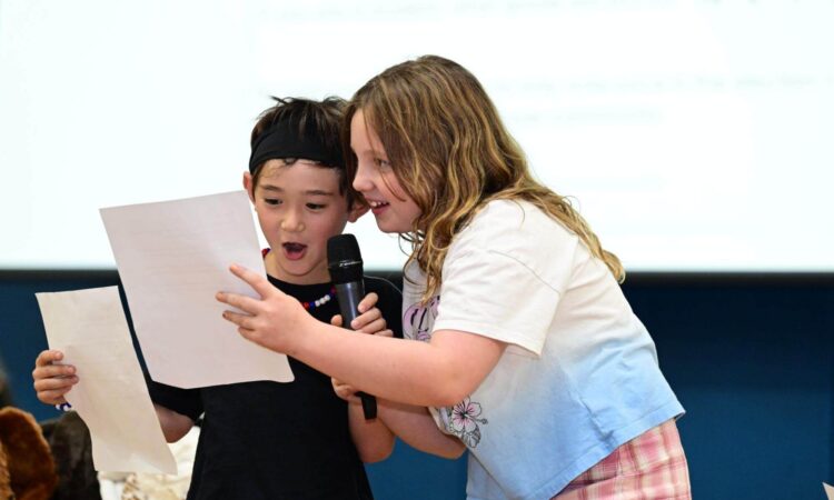 Two Ethical Culture Fieldston School Fieldston Lower students smile and share a piece of paper while presenting at an assembly.