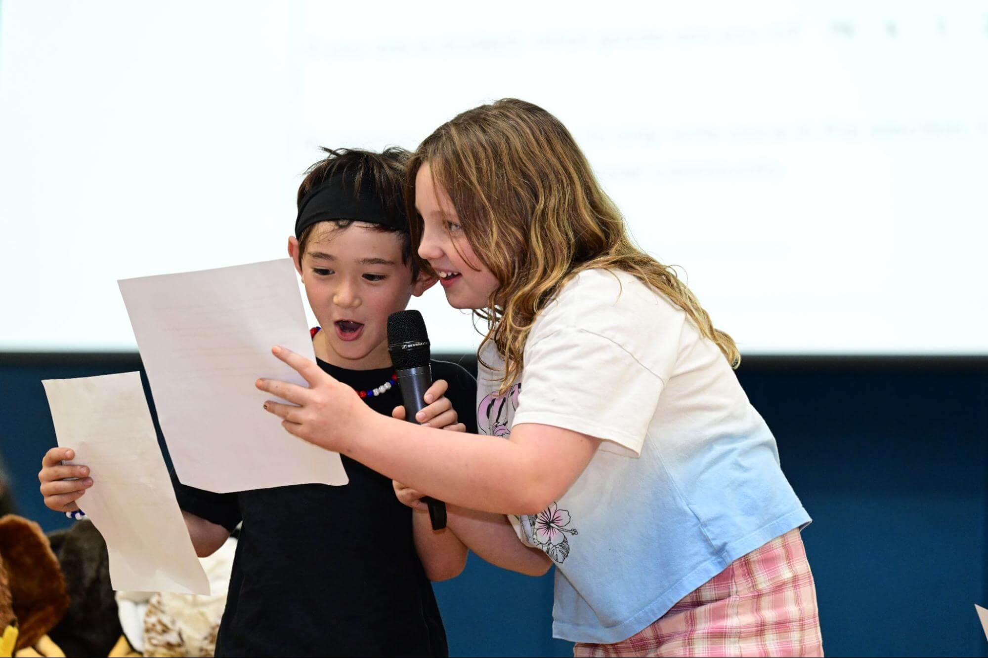 Two Ethical Culture Fieldston School Fieldston Lower students smile and share a piece of paper while presenting at an assembly.