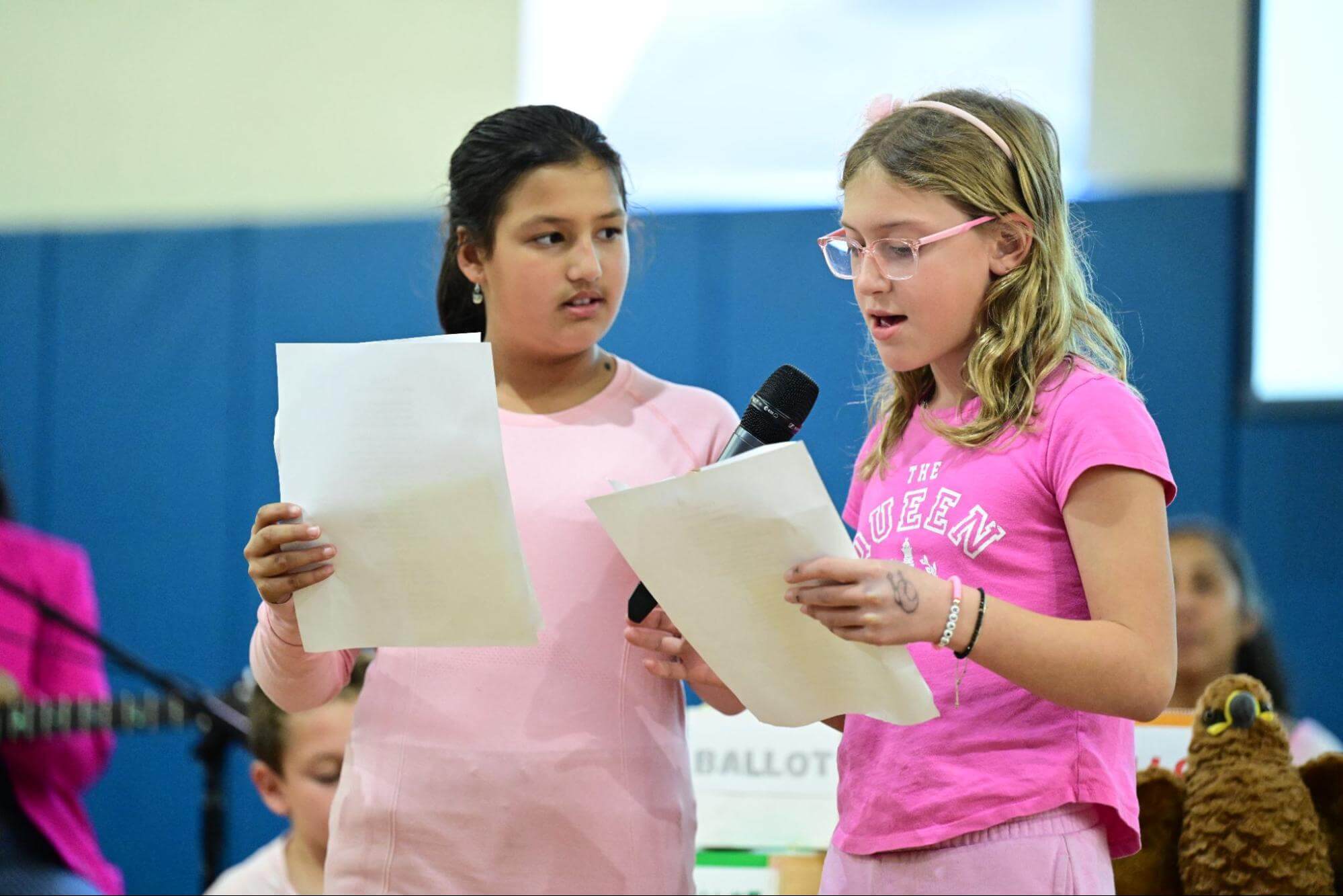 Two Ethical Culture Fieldston School Fieldston Lower students share a microphone while presenting at an assembly.