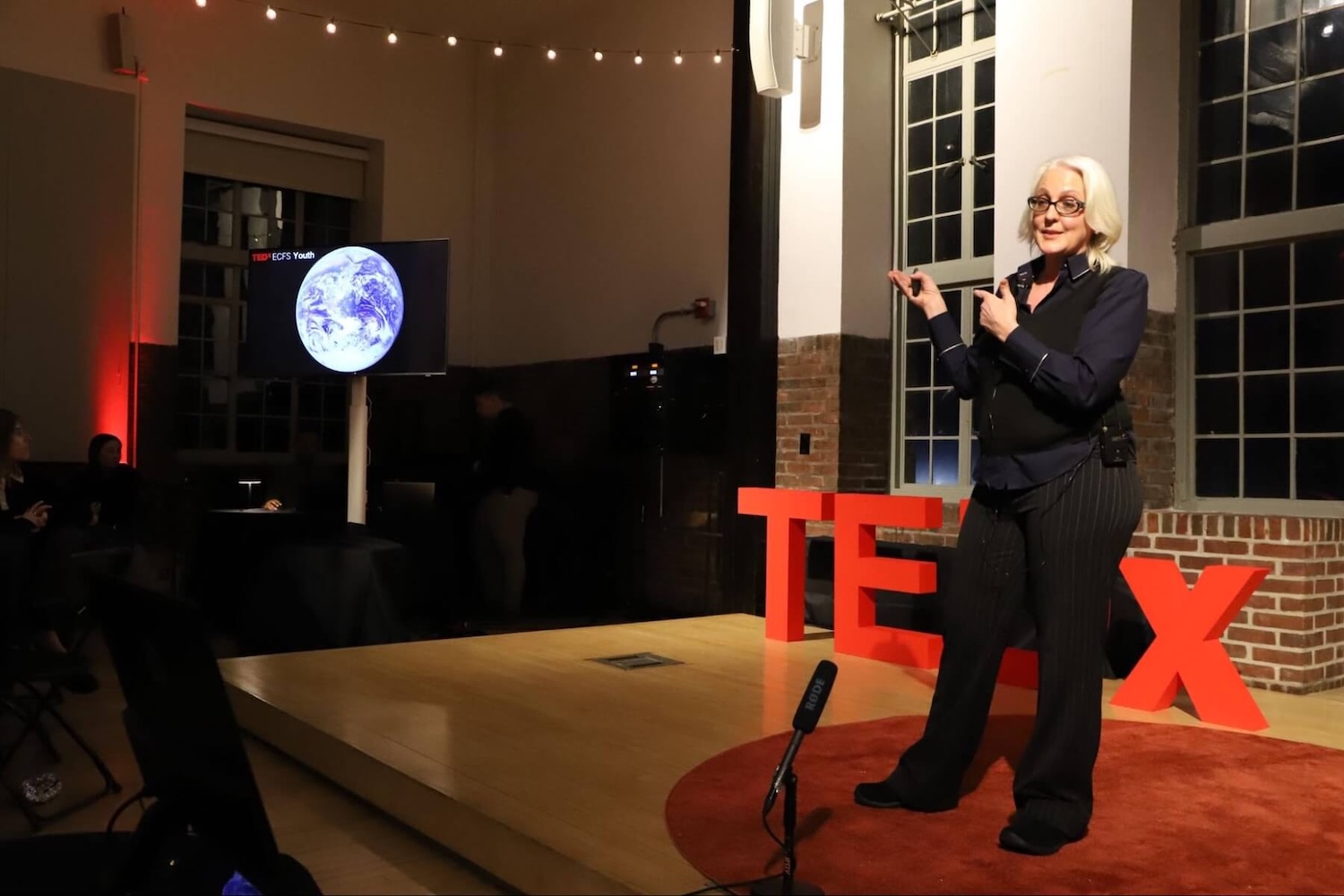 A Fieldston Upper science teacher speaks at the TEDxECFS event, gesturing to a TV displaying a picture of the Earth.