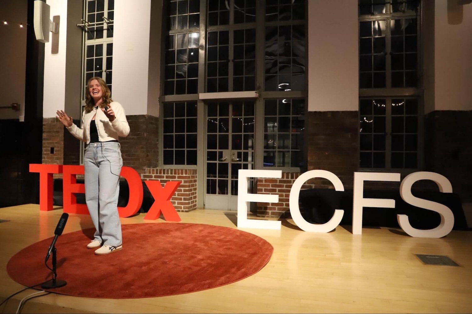 A smiling Fieldston Upper student speaks at the TEDxECFS event, standing in front of a red sign that reads "TEDx" and a white sign that reads "ECFS"