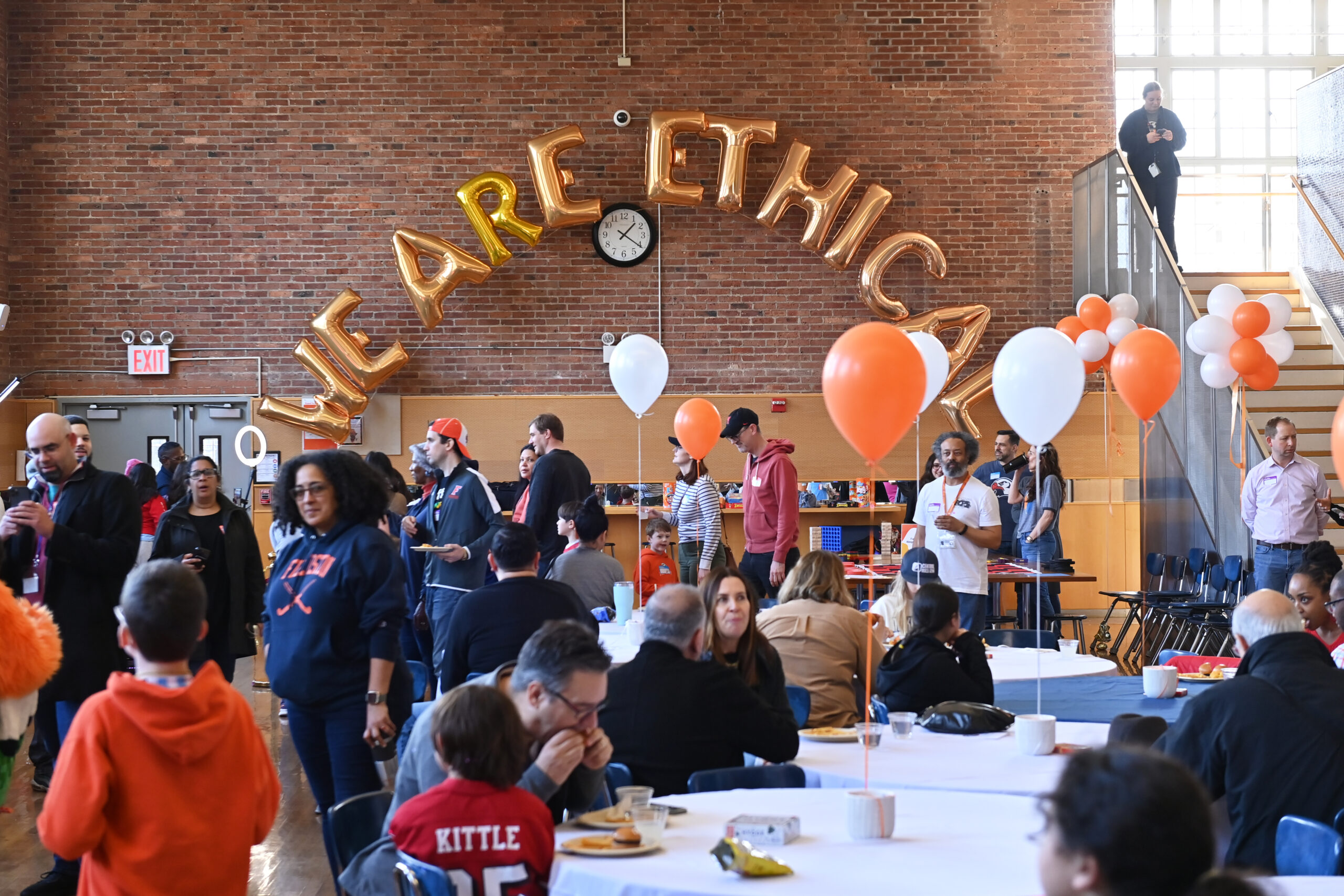 A large group of people sits and stands in the Ethical Culture Fieldston School dining room, surrounded by orange and white balloons and with "We Are Ethical" spelled out in gold balloons on the far wall.